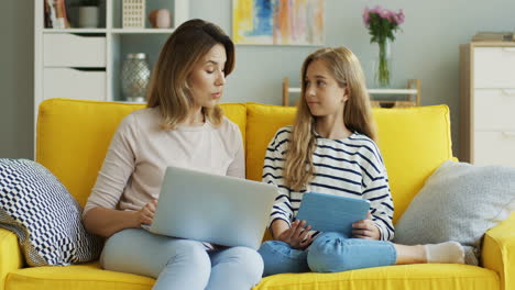 Cute-Caucasian-girl-talking-with-her-mother-while-sitting-on-the-yellow-sofa-with-laptop-computer-and-tablet-at-home.-Inside.